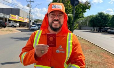 Homem sorridente em uniforme laranja de gari, óculos e barba, segura carteira da OAB na rua