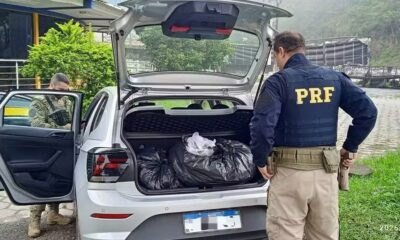 Dois policiais da PRF inspecionam porta-malas de carro prateado com grandes sacos