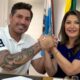Homem tatuado de polo branca com "3" e mulher de azul sorriem, mãos dadas, com bandeiras e "NEIRO".