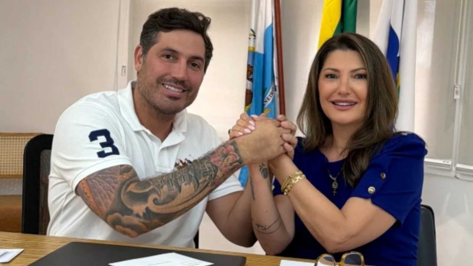 Homem tatuado de polo branca com "3" e mulher de azul sorriem, mãos dadas, com bandeiras e "NEIRO".