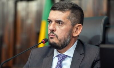 Homem de terno, camisa e gravata, com barba e microfone à frente, olha para a esquerda
