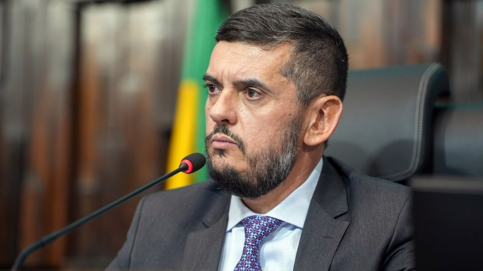 Homem de terno, camisa e gravata, com barba e microfone à frente, olha para a esquerda
