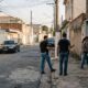 Homens de coletes e mulher em frente a portão de casa em rua