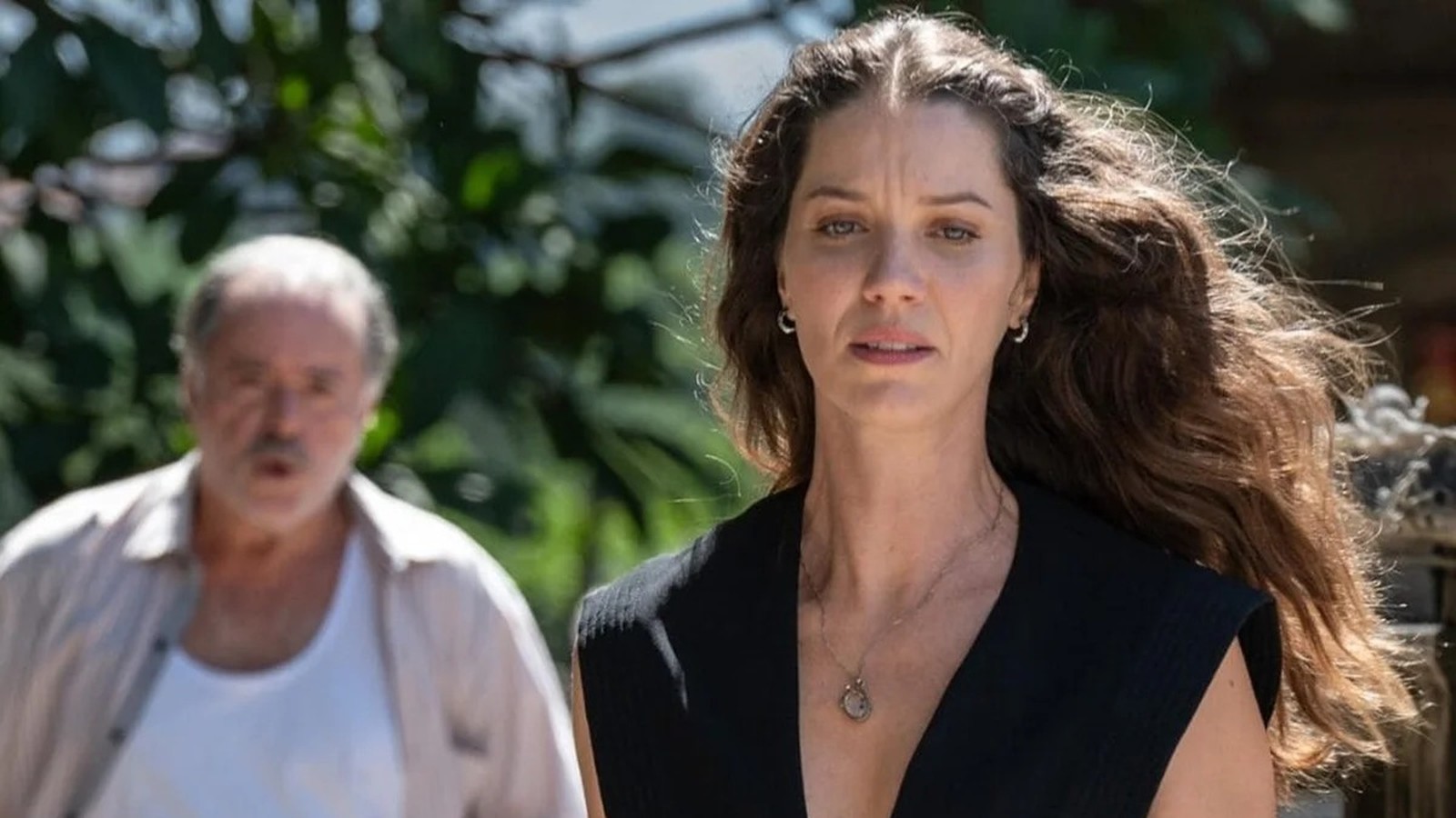 Nathalia Dill séria em primeiro plano com colar, homem desfocado atrás em cenário verde
