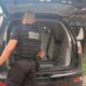 Polícia Federal officer, black uniform, next to open police SUV trunk; person visible inside