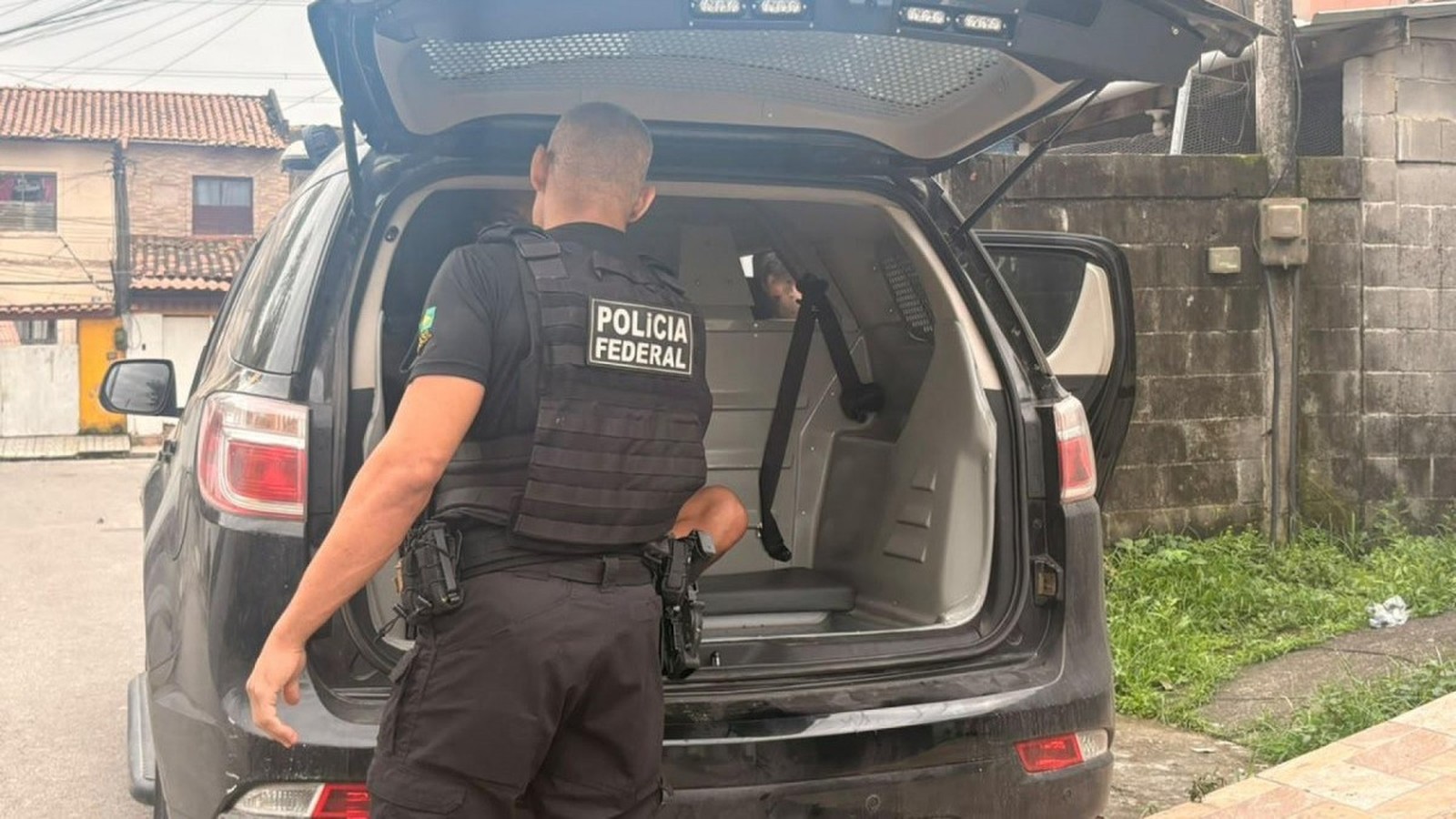 Polícia Federal officer, black uniform, next to open police SUV trunk; person visible inside