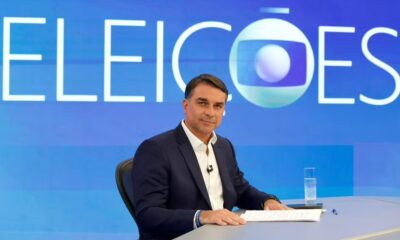 Homem de terno azul sentado, mesa com papéis, copo d'água. Fundo azul com "ELEIÇÕES" e logo Globo