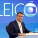 Homem de terno azul sentado, mesa com papéis, copo d'água. Fundo azul com "ELEIÇÕES" e logo Globo