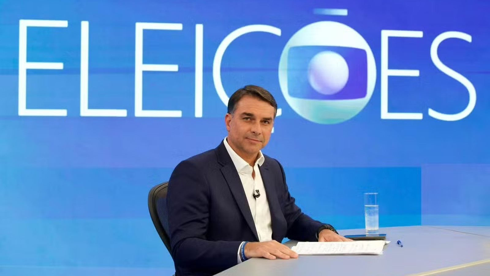 Homem de terno azul sentado, mesa com papéis, copo d'água. Fundo azul com "ELEIÇÕES" e logo Globo