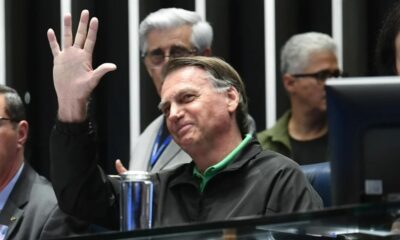 Jair Bolsonaro sorrindo e acenando com a mão direita, com pessoas desfocadas ao fundo