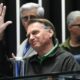 Jair Bolsonaro sorrindo e acenando com a mão direita, com pessoas desfocadas ao fundo
