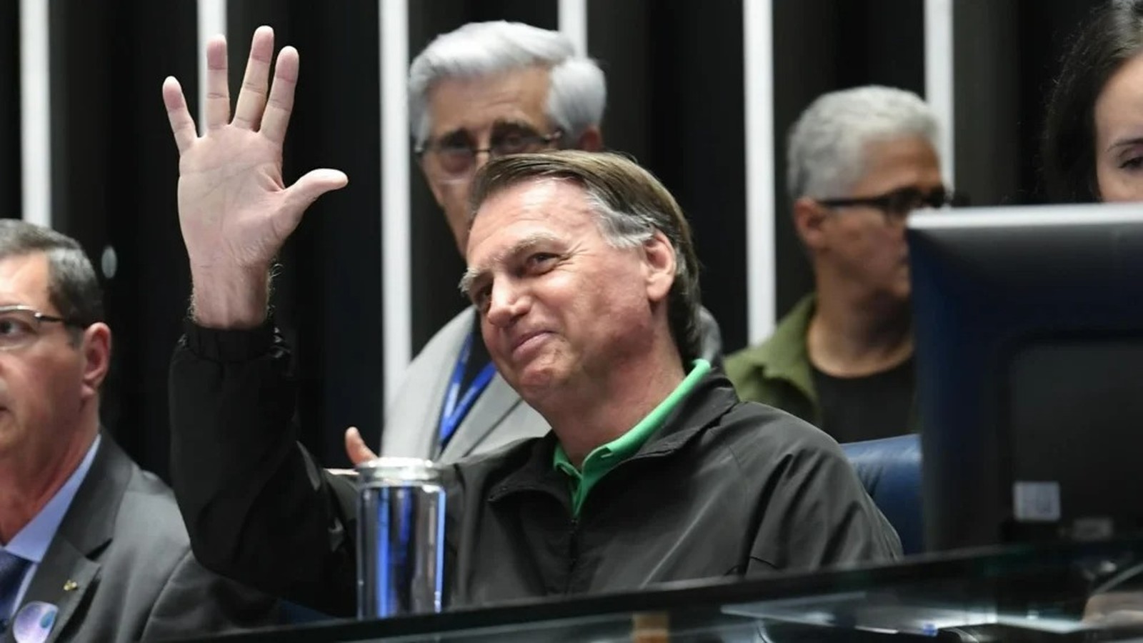 Jair Bolsonaro sorrindo e acenando com a mão direita, com pessoas desfocadas ao fundo