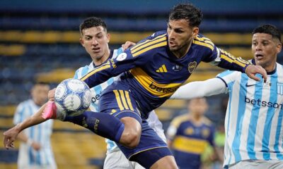 Três jogadores de futebol disputam uma bola em campo, um de uniforme azul e amarelo, dois de listras claras.