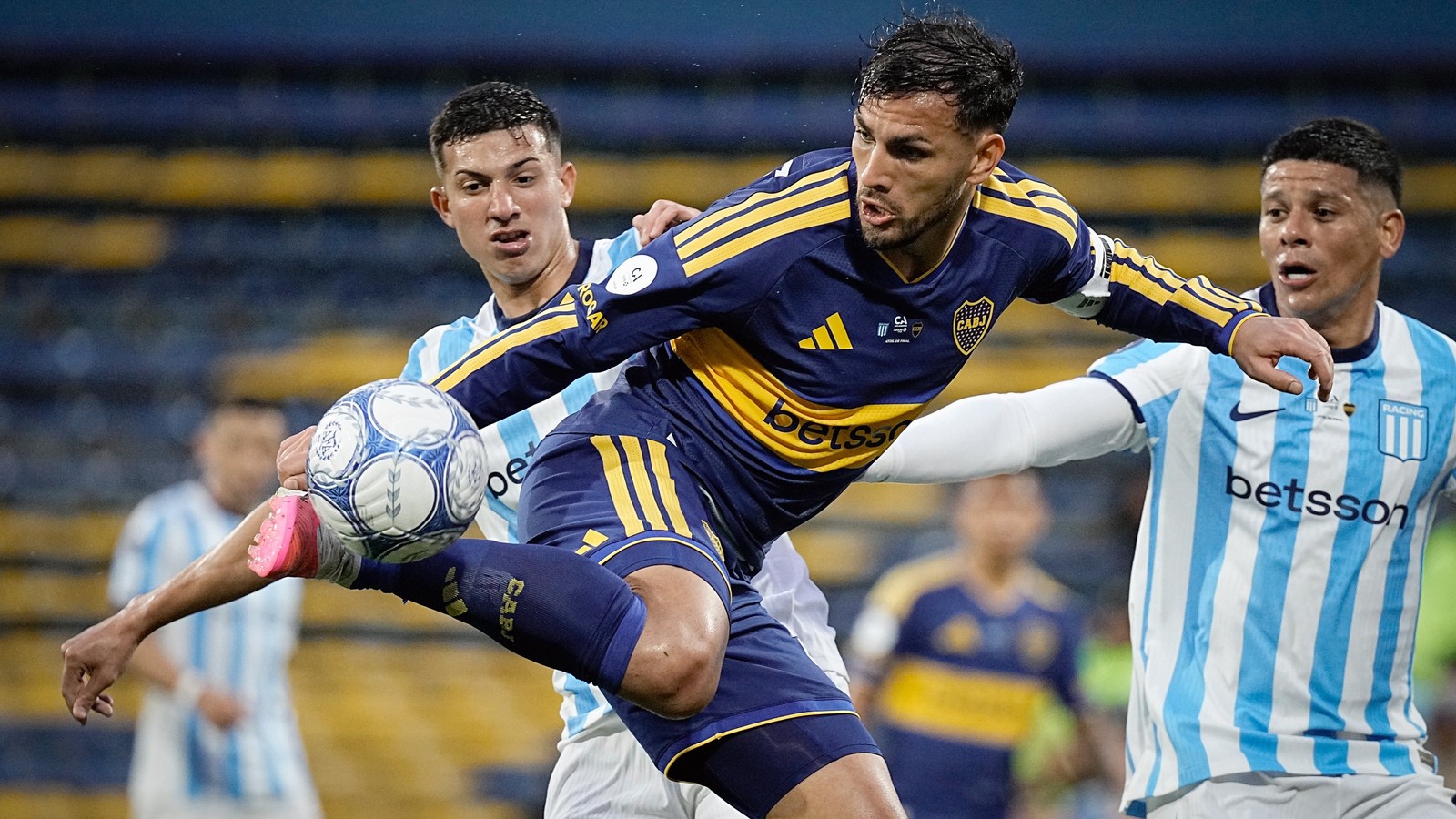Três jogadores de futebol disputam uma bola em campo, um de uniforme azul e amarelo, dois de listras claras.