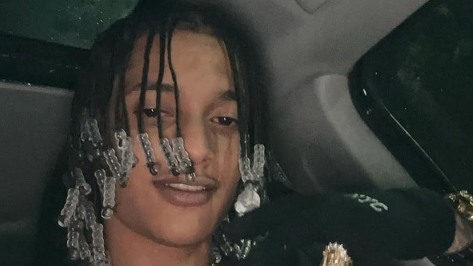 Oruam, homem jovem, dreadlocks com miçangas, sorri levemente dentro de um carro. Usa anéis