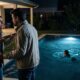 Mulher e homem discutem intensamente à beira da piscina, onde uma criança nada sozinha à noite.