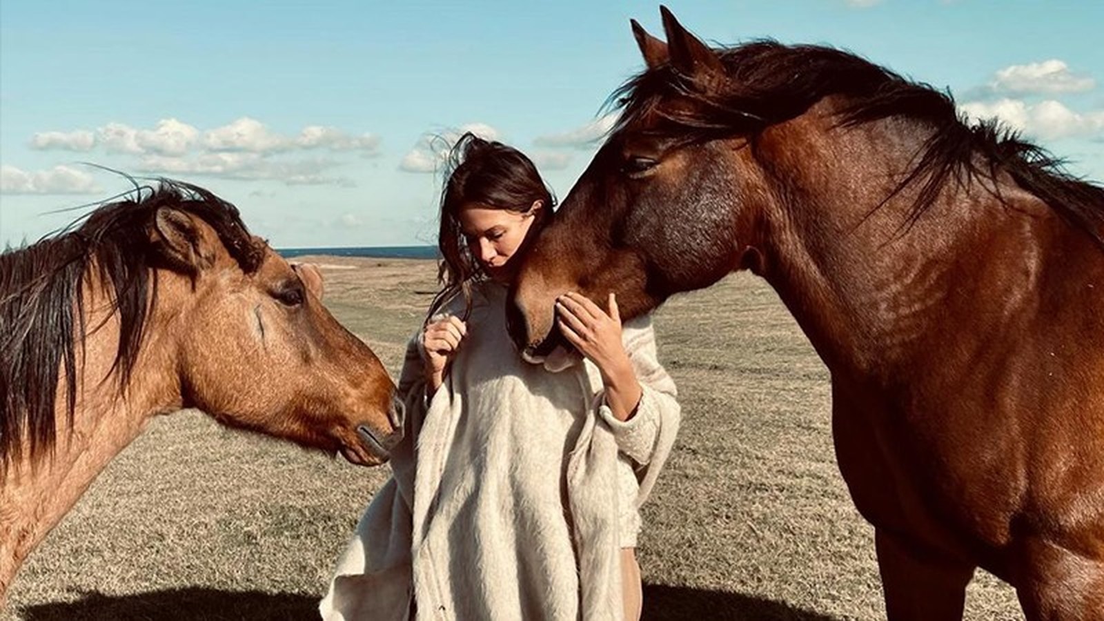 Mulher em poncho claro acaricia cavalo marrom, outro cavalo ao lado, em campo aberto