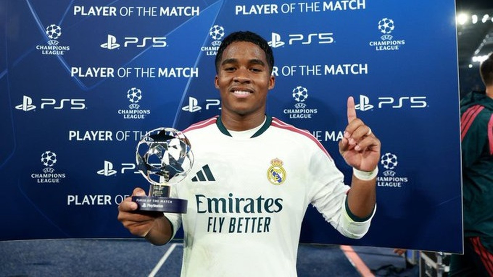Jogador Endrick sorri segurando troféu de Melhor em Campo da Champions League em fundo azul