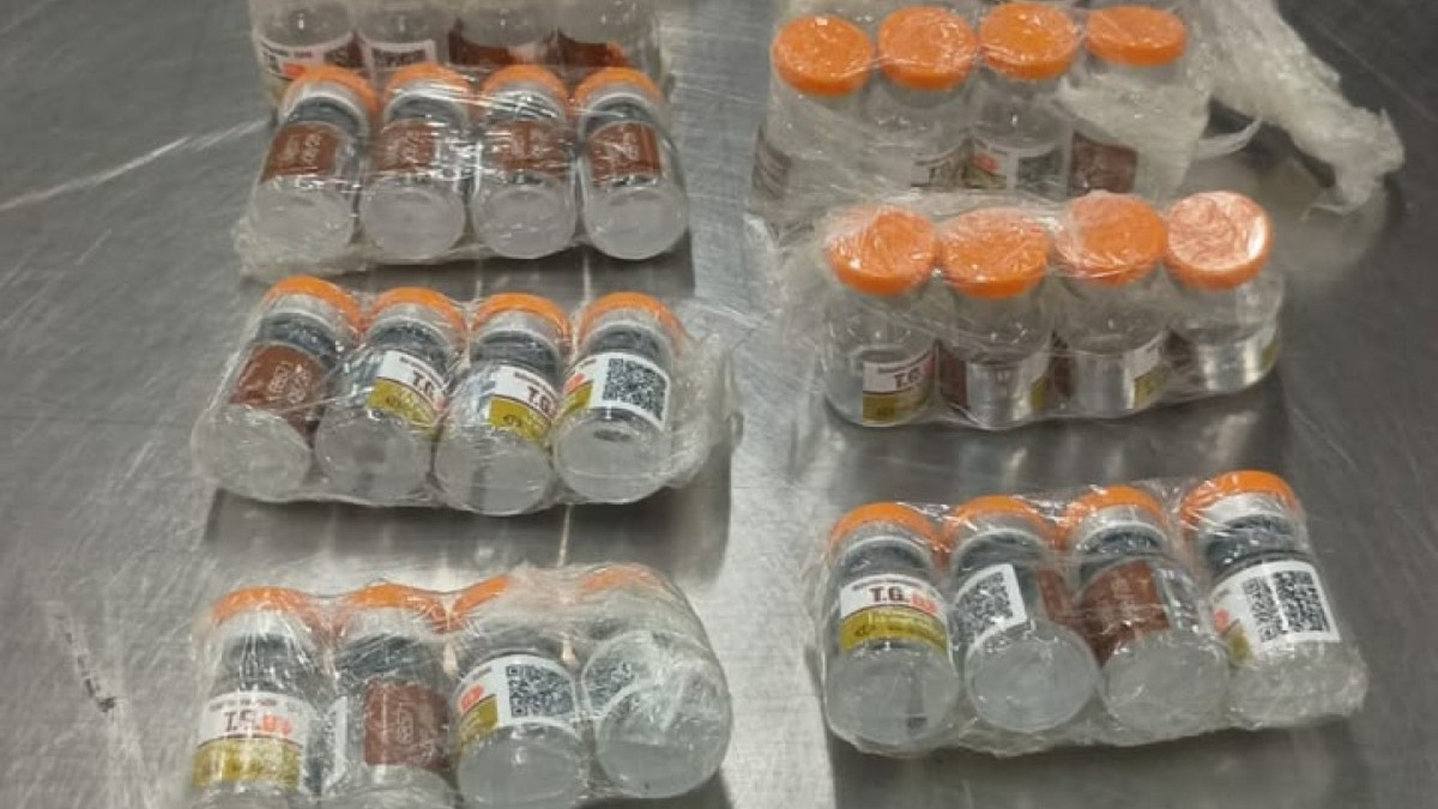 Muitos frascos de vidro com tampas laranja, alguns com rótulo 'T.G.' e códigos QR, embrulhados em plástico