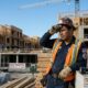 Homem trabalhador da construção civil secando o suor em canteiro com prédios e placa 'HELP WANTED'