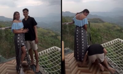 Jovem casal em varanda. Homem ajoelhado no chão de rede, mulher olha para baixo com montanhas ao fundo
