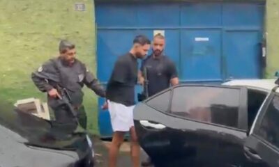 Policiais escoltam homem algemado de camisa preta e shorts brancos ao lado de carros e portão azul