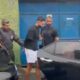 Policiais escoltam homem algemado de camisa preta e shorts brancos ao lado de carros e portão azul