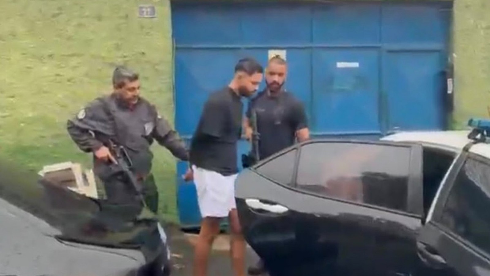 Policiais escoltam homem algemado de camisa preta e shorts brancos ao lado de carros e portão azul