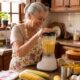 Mulher idosa sorri preparando bolo de fubá no liquidificador, com milho e ingredientes na bancada