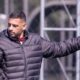 Homem de jaqueta escura e barba, apontando o dedo, em campo de futebol