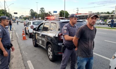 Homem com boné sendo detido por policial militar ao lado de viaturas e cones na rua