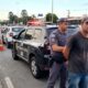 Homem com boné sendo detido por policial militar ao lado de viaturas e cones na rua