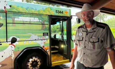 Homem de chapéu e uniforme sorri ao lado de ônibus verde da Disney com arte de Dálmatas e número 5344