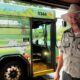 Homem de chapéu e uniforme sorri ao lado de ônibus verde da Disney com arte de Dálmatas e número 5344