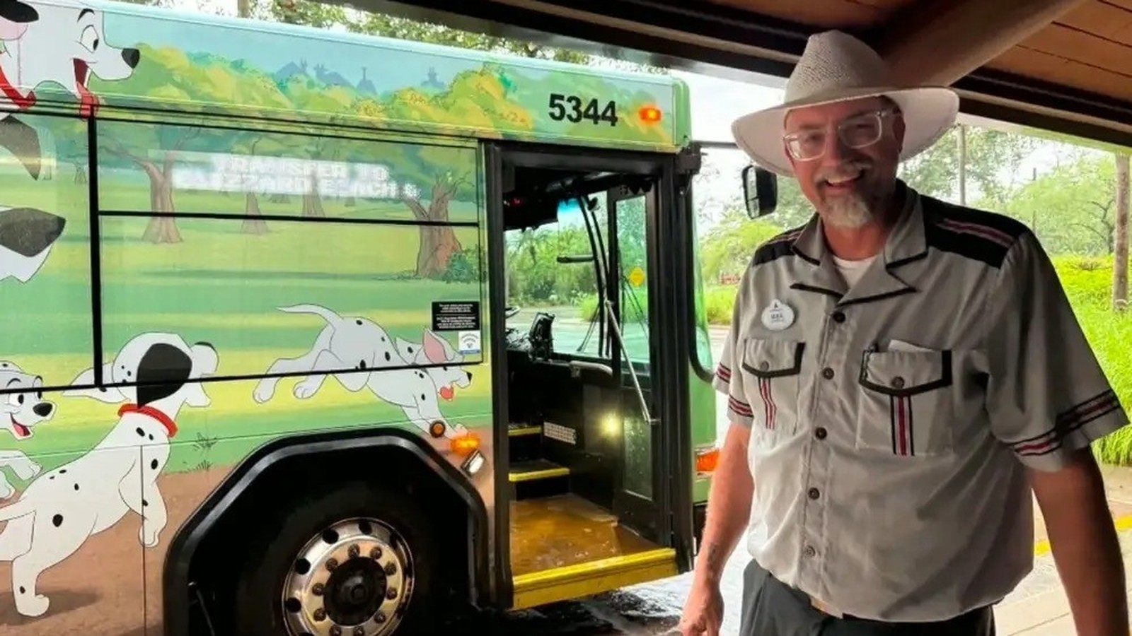 Homem de chapéu e uniforme sorri ao lado de ônibus verde da Disney com arte de Dálmatas e número 5344