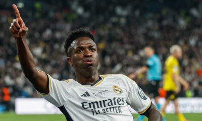 Vini Jr. de camisa branca do Real Madrid, apontando para cima, em campo, com torcida ao fundo
