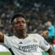 Vini Jr. de camisa branca do Real Madrid, apontando para cima, em campo, com torcida ao fundo