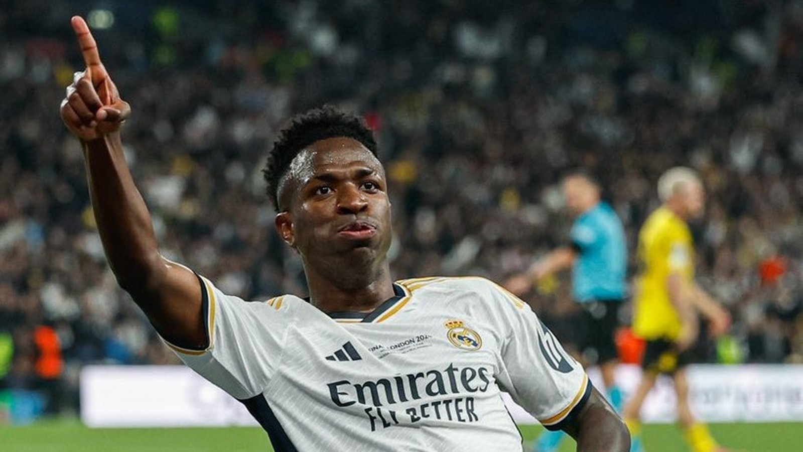 Vini Jr. de camisa branca do Real Madrid, apontando para cima, em campo, com torcida ao fundo