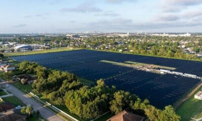 Vista aérea de usina solar com painéis pretos em área urbana, casas e skyline ao fundo