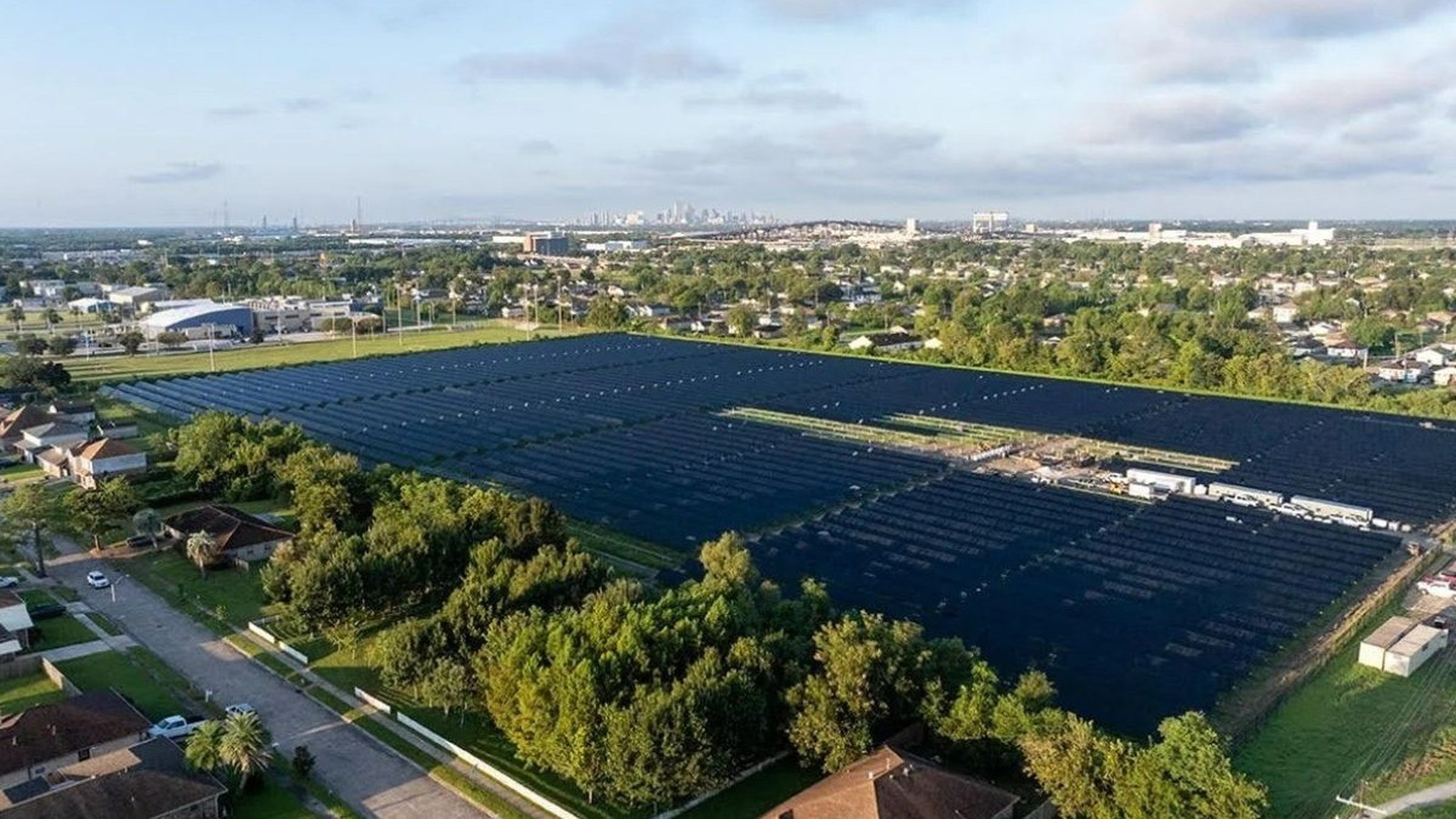 Vista aérea de usina solar com painéis pretos em área urbana, casas e skyline ao fundo