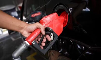 Mão inserindo bico de bomba de combustível em tanque de carro preto, escrito GASOLINA