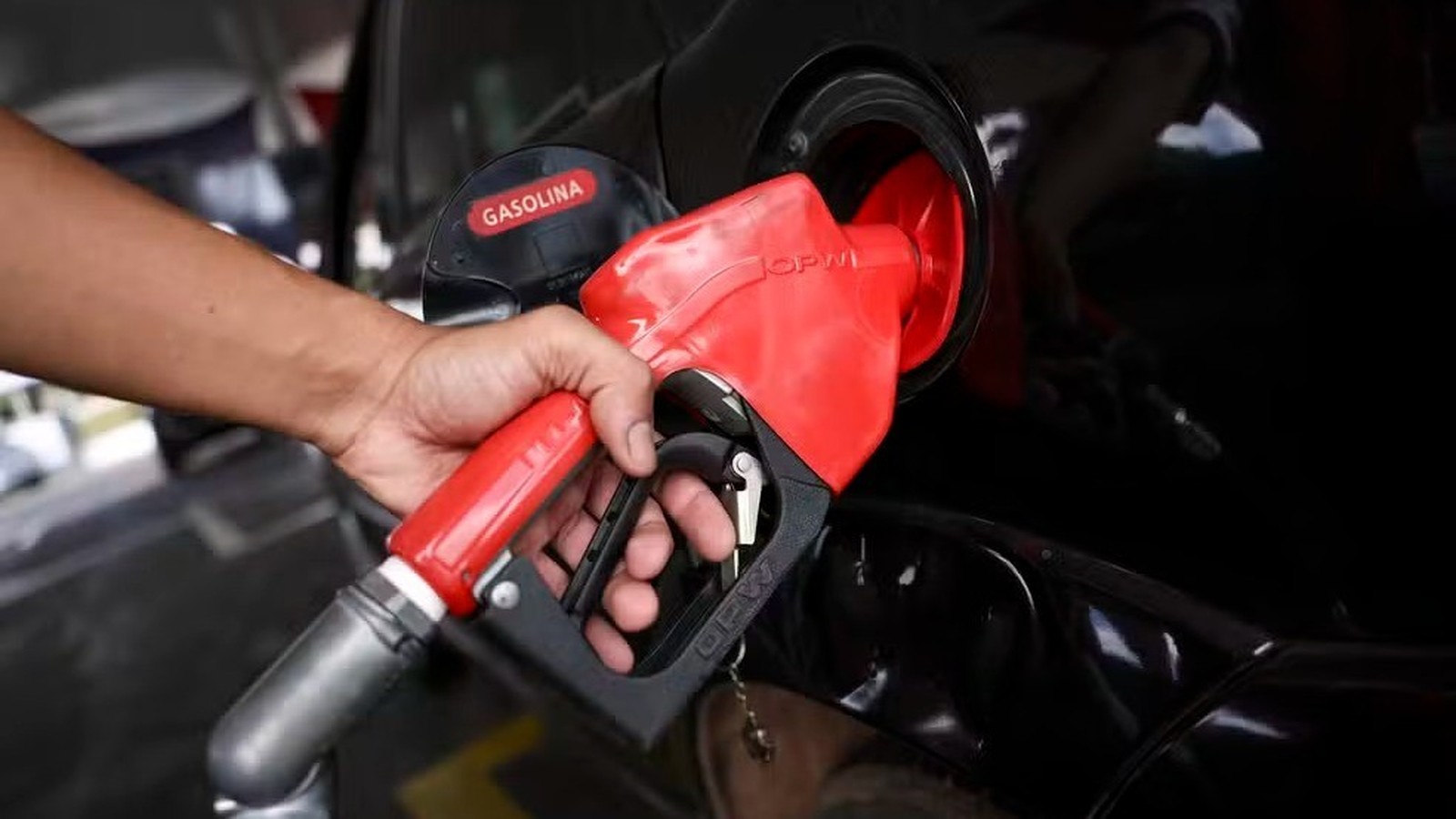 Mão inserindo bico de bomba de combustível em tanque de carro preto, escrito GASOLINA