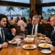 Quatro homens de terno em restaurante. Um deles com rosto zangado e punhos cerrados