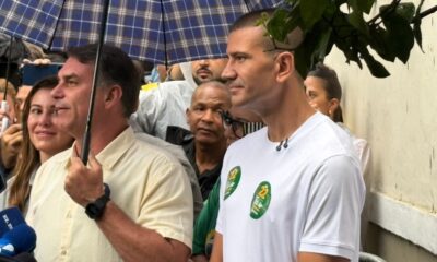 Flávio Bolsonaro, com guarda-chuva, entre apoiadores, incluindo homem com camiseta branca e broches "22"