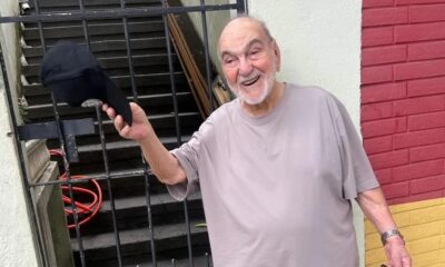 Lima Duarte, idoso, sorrindo e segurando boné preto, com portão e parede ao fundo.
