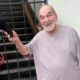 Lima Duarte, idoso, sorrindo e segurando boné preto, com portão e parede ao fundo.