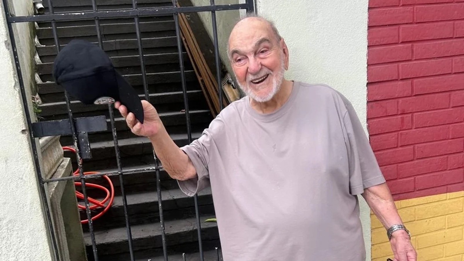 Lima Duarte, idoso, sorrindo e segurando boné preto, com portão e parede ao fundo.