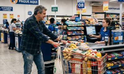 Homem sorridente empurra carrinho de compras lotado para caixa de supermercado com atendente mulher