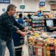 Homem sorridente empurra carrinho de compras lotado para caixa de supermercado com atendente mulher
