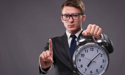 Homem de terno, óculos, gravata, segura despertador grande e levanta o dedo indicador esquerdo